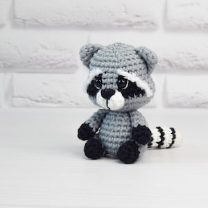 May include: A small crocheted raccoon plush toy with black eyes, a white nose, and a black and white striped tail. The toy is sitting on a white surface.