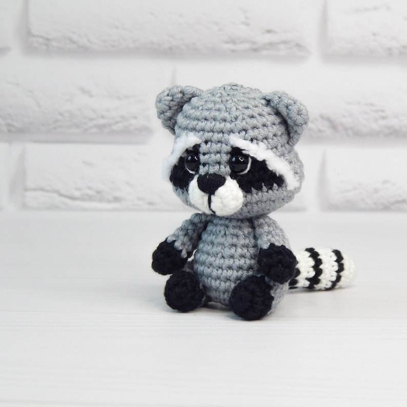 May include: A small crocheted raccoon plush toy with black eyes, a white nose, and a black and white striped tail. The toy is sitting on a white surface.
