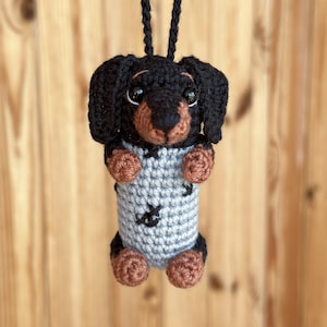 May include: A handmade crochet dachshund dog ornament. The dog has a black body, brown paws and snout, and large, expressive eyes. It wears a gray sweater with black accents and is suspended by a black cord. A charming, whimsical decoration.