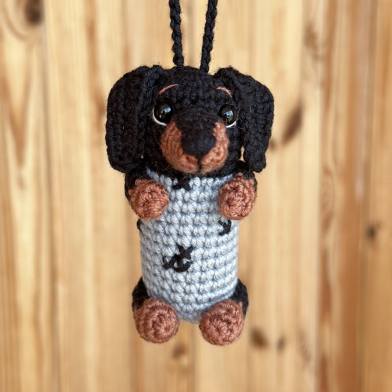 May include: A handmade crochet dachshund dog ornament. The dog has a black body, brown paws and snout, and large, expressive eyes. It wears a gray sweater with black accents and is suspended by a black cord. A charming, whimsical decoration.