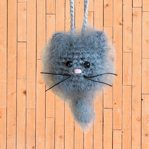 May include: A gray crocheted cat-shaped bag with black whiskers and pink nose. The bag is hanging from a wooden background.