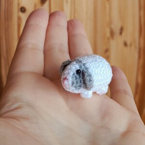 Pode incluir: Um pequeno brinquedo de cobaia branco e cinza de croch&ecirc; com nariz rosa e olhos pretos.