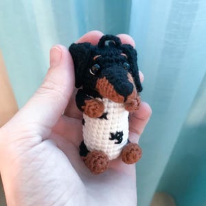 May include: A handmade crochet dachshund dog with black, brown, and white details. The dog has a black head and ears, brown paws, and a white body with black star and heart accents. It has a small loop for hanging.