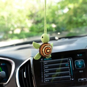 Puede incluir: Una tortuga verde de crochet con una concha marr&oacute;n, naranja y amarilla cuelga del espejo retrovisor de un coche.