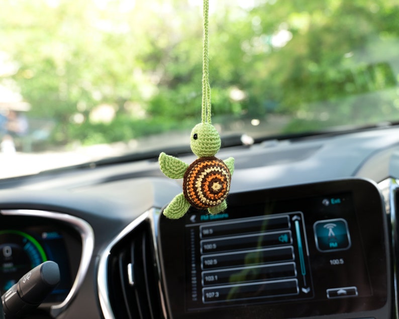Puede incluir: Una tortuga verde de crochet con una concha marr&oacute;n, naranja y amarilla cuelga del espejo retrovisor de un coche.