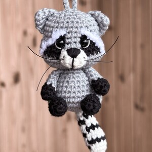 May include: A crocheted raccoon ornament with a gray body, black eyes, white nose, and black and white striped tail.