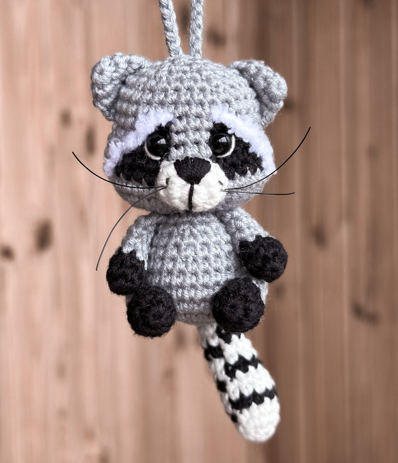 May include: A crocheted raccoon ornament with a gray body, black eyes, white nose, and black and white striped tail.