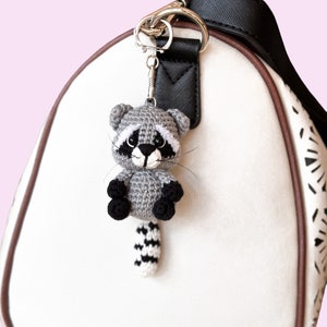 May include: A gray crocheted raccoon keychain with black eyes and a black and white striped tail. The keychain is attached to a silver key ring and is hanging from a black purse strap.
