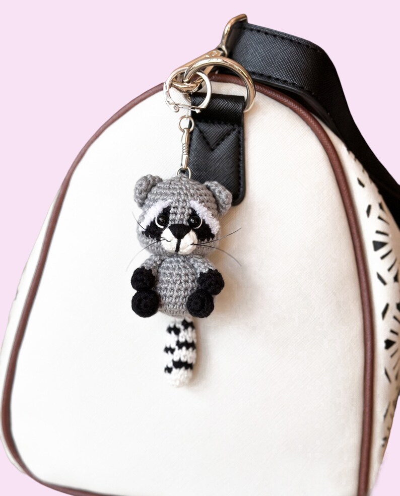 May include: A gray crocheted raccoon keychain with black eyes and a black and white striped tail. The keychain is attached to a silver key ring and is hanging from a black purse strap.