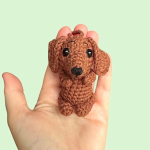 May include: A small, handmade crochet dachshund dog in shades of brown. The dog has black eyes and a black nose. It is held in a person's hand against a light green background. The dog is a miniature version of the breed.