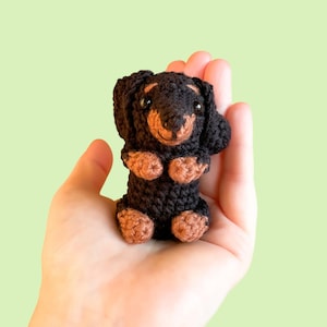 May include: A handmade crochet dachshund dog in black and brown. The small dog has a long body, short legs, and floppy ears. The dog is held in a hand against a light green background.