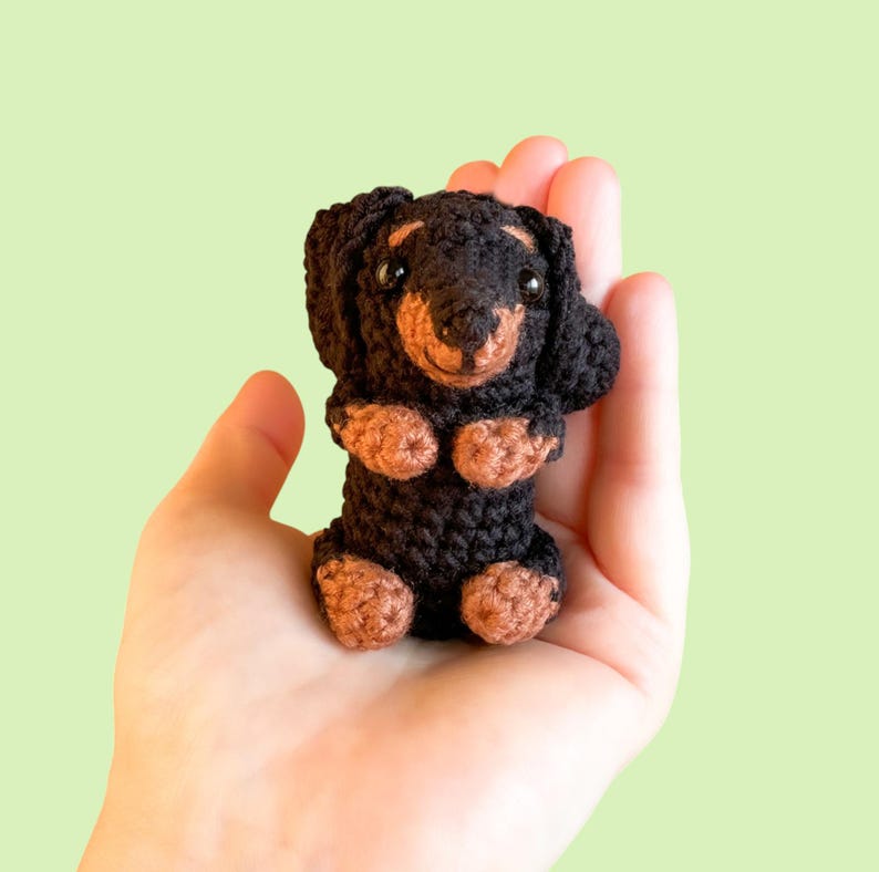 May include: A handmade crochet dachshund dog in black and brown. The small dog has a long body, short legs, and floppy ears. The dog is held in a hand against a light green background.