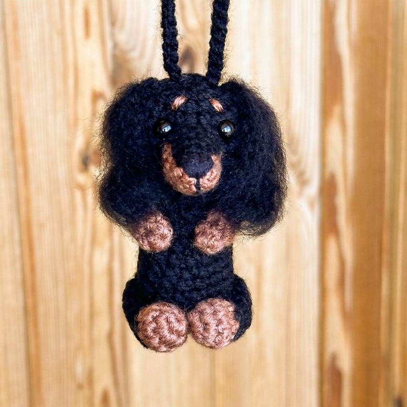 May include: A handmade black and brown crochet dachshund dog with a black cord for hanging. The dog has a fluffy head, small black eyes, and brown paws and snout. The background is a light brown wooden surface.