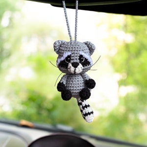 May include: A gray crocheted raccoon hanging from a rearview mirror. The raccoon has black eyes, a white nose, and a black and white striped tail.