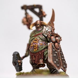 May include: A green and brown painted miniature figure of a grotesque, fleshy creature with a large hammer and shield. The figure is wearing a horned helmet and has a large, fleshy belly covered in yellow and orange spots.