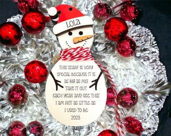 May include: A wooden snowman Christmas ornament with a red and white hat and scarf. The ornament has the name "Lola" on the hat and a message about the scarf. The ornament is surrounded by red and silver Christmas decorations.