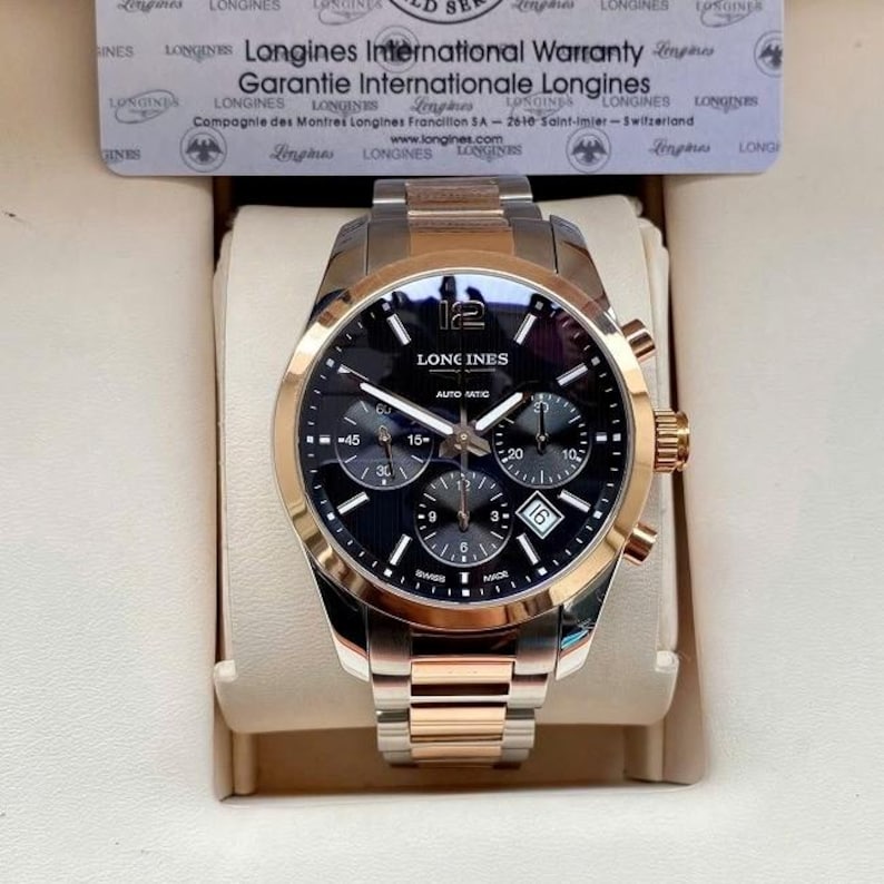 May include: A Longines automatic watch with a black dial, gold-tone bezel, and stainless steel band. The watch face displays the Longines logo, sub-dials, and a date window. The watch is presented in a display box with a warranty card.