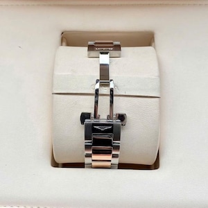 May include: A close-up of a watch clasp, featuring a silver and rose gold band. The clasp is secured, showcasing the Longines logo. The watch is set against a cream-colored background, suggesting a presentation box.