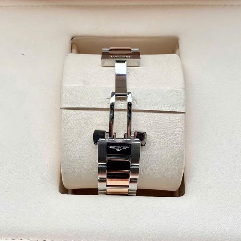 May include: A close-up of a watch clasp, featuring a silver and rose gold band. The clasp is secured, showcasing the Longines logo. The watch is set against a cream-colored background, suggesting a presentation box.