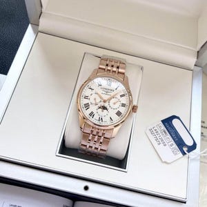 May include: A rose gold Longines automatic watch with a white dial, Roman numeral hour markers, and a moon phase indicator. The watch has a matching rose gold bracelet and is presented in a white box. A tag with the Longines logo and model number is attached.