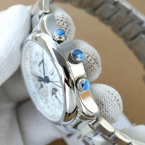 Op de afbeelding: Een zilveren polshorloge met een witte wijzerplaat en blauwe wijzers. Het horloge heeft een zilveren band en drie blauwe accenten aan de zijkant. Het horloge wordt vastgehouden door een gehandschoende hand.