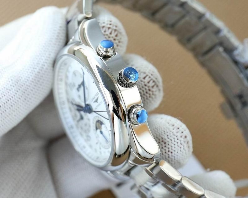 Op de afbeelding: Een zilveren polshorloge met een witte wijzerplaat en blauwe wijzers. Het horloge heeft een zilveren band en drie blauwe accenten aan de zijkant. Het horloge wordt vastgehouden door een gehandschoende hand.