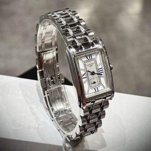 Könnte beinhalten: Eine rechteckige Armbanduhr in Silber mit weißem Zifferblatt und schwarzen römischen Ziffern. Die Uhr hat ein Gliederarmband, und der Markenname "Longines" ist auf dem Zifferblatt zu sehen. Die Uhr wird auf einem transparenten Ständer präsentiert.