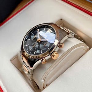 May include: A luxury chronograph watch with a black dial, gold accents, and a stainless steel band. The watch features multiple sub-dials and a date display. The watch is presented in a white and red box.