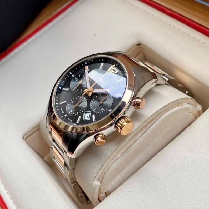 May include: A luxury chronograph watch with a black dial, gold accents, and a stainless steel band. The watch features multiple sub-dials and a date display. The watch is presented in a white and red box.
