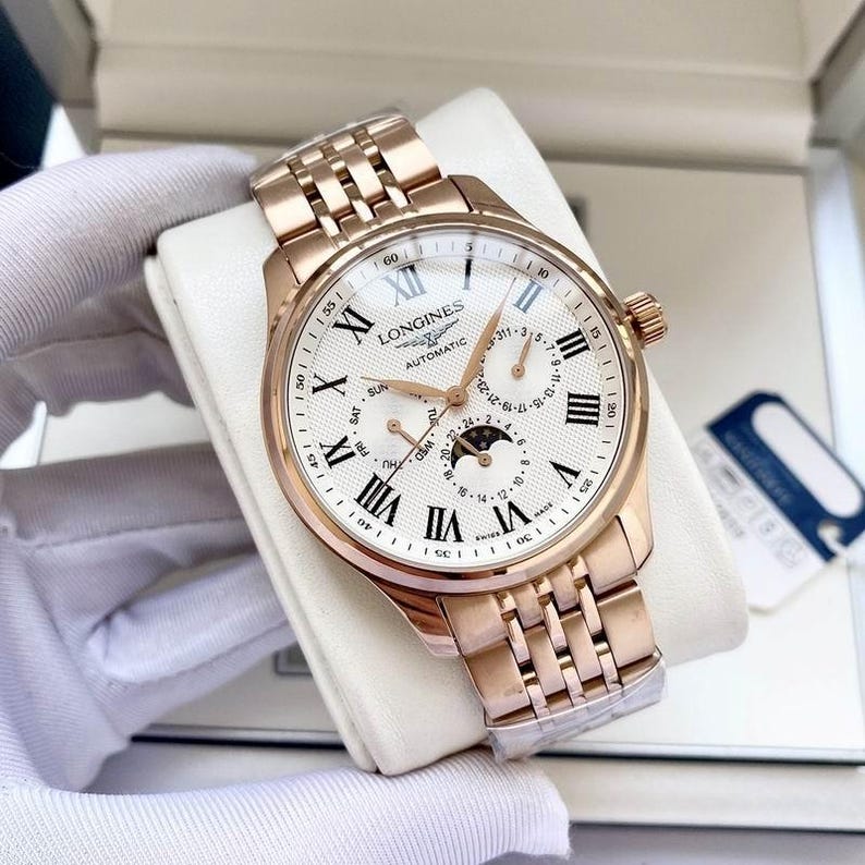 May include: A rose gold Longines automatic watch with a white dial, featuring Roman numeral hour markers and multiple sub-dials. The watch has a matching rose gold bracelet and a crown on the right side. The watch is displayed on a white cushion.