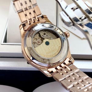 May include: Rose gold wristwatch with a transparent back, revealing the internal mechanism. The watch has a metal strap and a round face. The text "PERPETUAL CALENDAR" and "SWISS MADE" are visible.