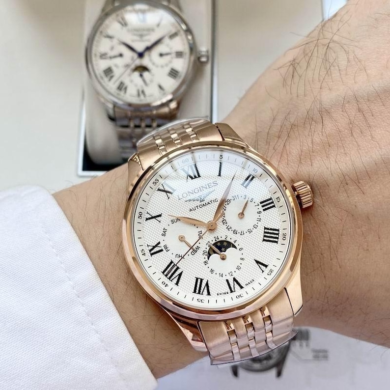 May include: A rose gold Longines automatic watch with a white face, Roman numeral hour markers, and a moon phase indicator. The watch has a stainless steel band and is displayed on a wrist. Another watch is visible in the background.