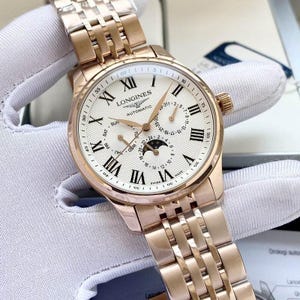 May include: A rose gold Longines automatic watch with a white face and Roman numeral hour markers. The watch features a date display, day of the week, and moon phase indicator. The watch band is also rose gold.