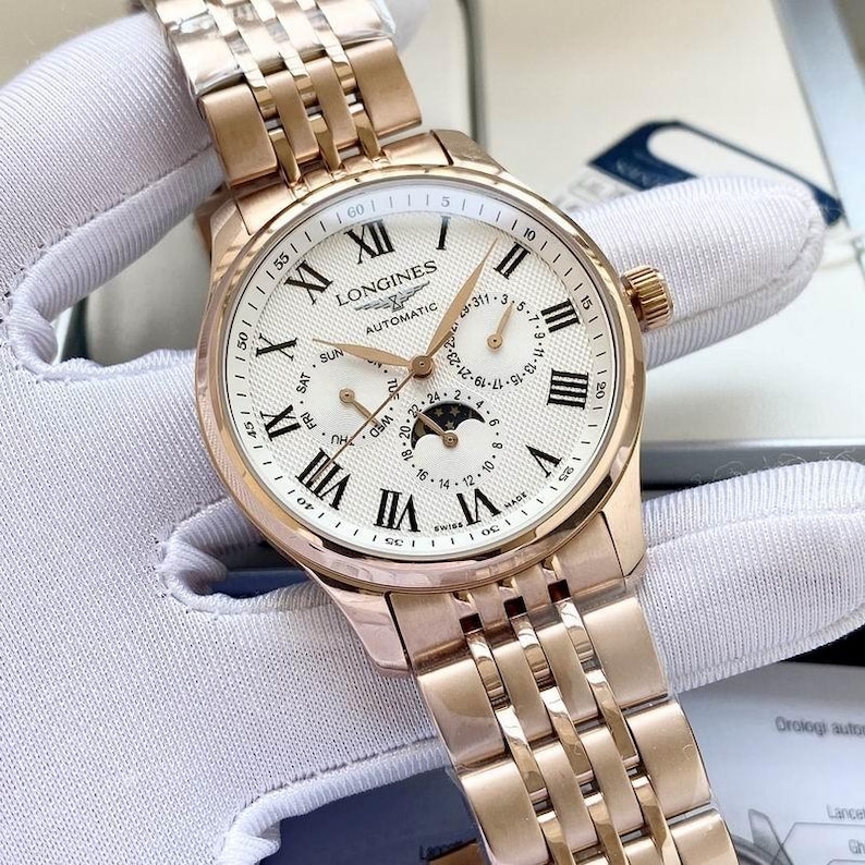 May include: A rose gold Longines automatic watch with a white face and Roman numeral hour markers. The watch features a date display, day of the week, and moon phase indicator. The watch band is also rose gold.