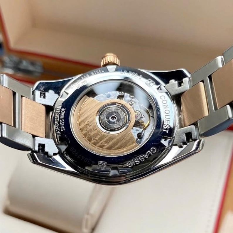 May include: Close-up of a stainless steel and rose gold watch. The watch features a clear back, showcasing the intricate inner workings. The band is a combination of silver and rose gold links. The watch is labeled "CONQUEST CLASSIC" and "SWISS MADE".