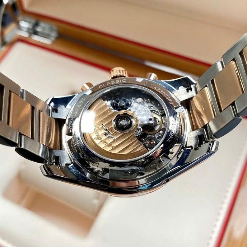 May include: A close-up of a luxury wristwatch with a silver and gold-tone metal band. The watch face is visible, showcasing the intricate internal mechanisms. The watch has a classic design with the word "CLASSIC" on the top.