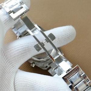 Op de afbeelding: Close-up van een zilveren roestvrijstalen horlogebandsluiting. De sluiting is open en onthult de binnenkant. Het woord "LONGINES" is op de band gegraveerd. De horlogeband wordt vastgehouden door een gehandschoende hand.