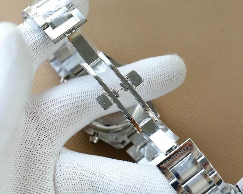 Op de afbeelding: Close-up van een zilveren roestvrijstalen horlogebandsluiting. De sluiting is open en onthult de binnenkant. Het woord "LONGINES" is op de band gegraveerd. De horlogeband wordt vastgehouden door een gehandschoende hand.