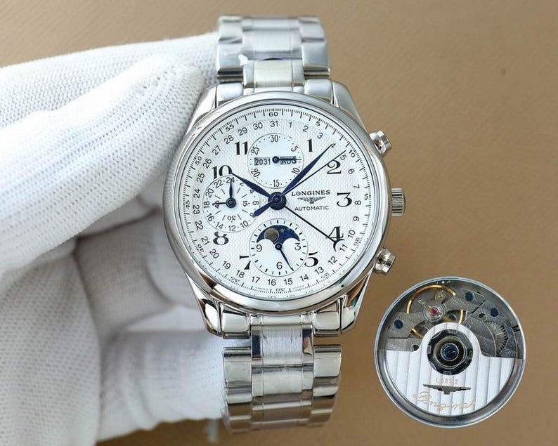 Op de afbeelding: Een zilverkleurig Longines automatisch horloge met een witte wijzerplaat, meerdere subwijzerplaten en een datumweergave. Het horloge heeft een roestvrijstalen band en een aparte weergave van het binnenwerk.