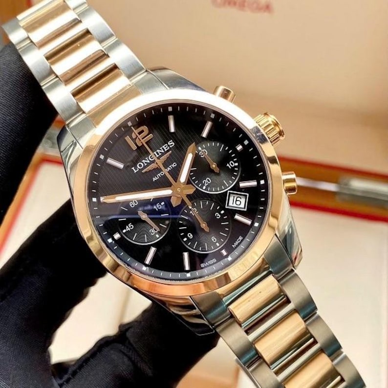 May include: A Longines automatic watch with a black dial, gold accents, and a stainless steel and gold-tone bracelet. The watch features chronograph subdials and a date window at the 3 o'clock position. The watch is Swiss made.