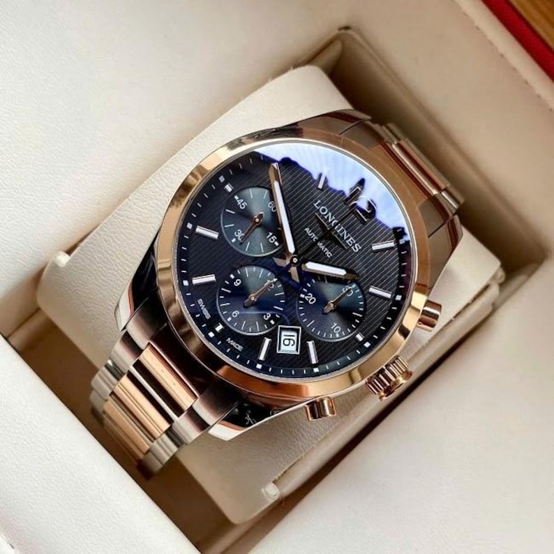 May include: A luxury Longines automatic watch with a black dial, gold-tone bezel, and a stainless steel and gold-tone bracelet. The watch features multiple subdials and a date display at the 6 o'clock position. The watch is presented in a cream-colored box.