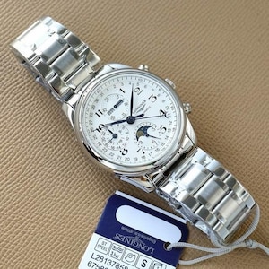 Op de afbeelding: Een zilverkleurig Longines automatisch horloge met een witte wijzerplaat, met meerdere subwijzerplaten en een datumweergave. Het horloge heeft een roestvrijstalen band en wordt gepresenteerd met een label. De wijzerplaat bevat de tekst "LONGINES".