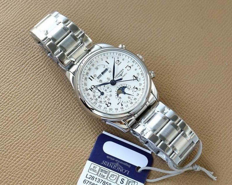 Op de afbeelding: Een zilverkleurig Longines automatisch horloge met een witte wijzerplaat, met meerdere subwijzerplaten en een datumweergave. Het horloge heeft een roestvrijstalen band en wordt gepresenteerd met een label. De wijzerplaat bevat de tekst "LONGINES".