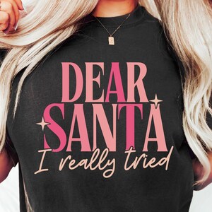 Könnte beinhalten: Ein schwarzes T-Shirt mit dem Text "Dear Santa I really tried" in rosa Schrift. Der Text ist in einer fetten, klobigen Schriftart.