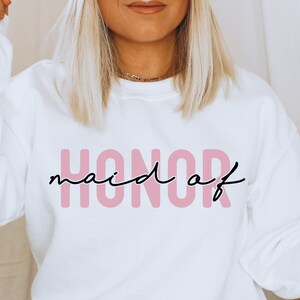May include: White sweatshirt with the text "maid of HONOR" in pink letters. The text is in a modern, handwritten font.