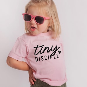 May include: A young child wearing pink sunglasses and a pink t-shirt with the text "tiny disciple" in black lettering. The child is standing in front of a white background.