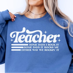 May include: A blue sweatshirt with the word "Teacher" in a white, retro font. Below the word are the phrases "SOME DAYS I ROCK IT", "SOME DAYS IT ROCKS ME", and "EITHER WAY WE ROCKIN' IT" in white. The sweatshirt is a solid blue colour.