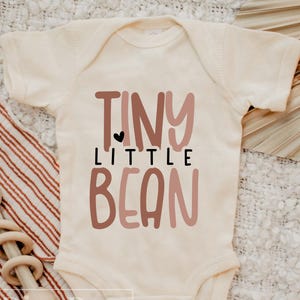 Könnte beinhalten: Ein weißer Baby-Strampler mit dem Text "TINY LITTLE BEAN" in rosa und braunen Buchstaben. Ein kleines schwarzes Herz befindet sich über dem Wort "LITTLE".