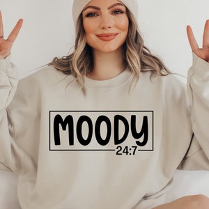 May include: A beige sweatshirt with the text "MOODY 24:7" printed in black. The model is wearing a beige beanie and is making a peace sign with both hands.