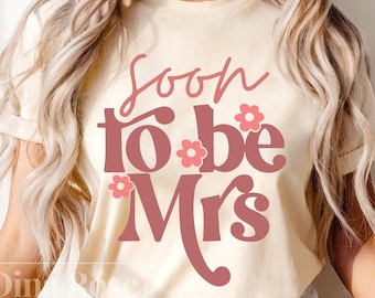 Puede incluir: Camiseta color crema con la frase "Soon to be Mrs" en una fuente retro y detalles florales. El texto es de color rosa apagado, ideal para una despedida de soltera o una fiesta de bodas.